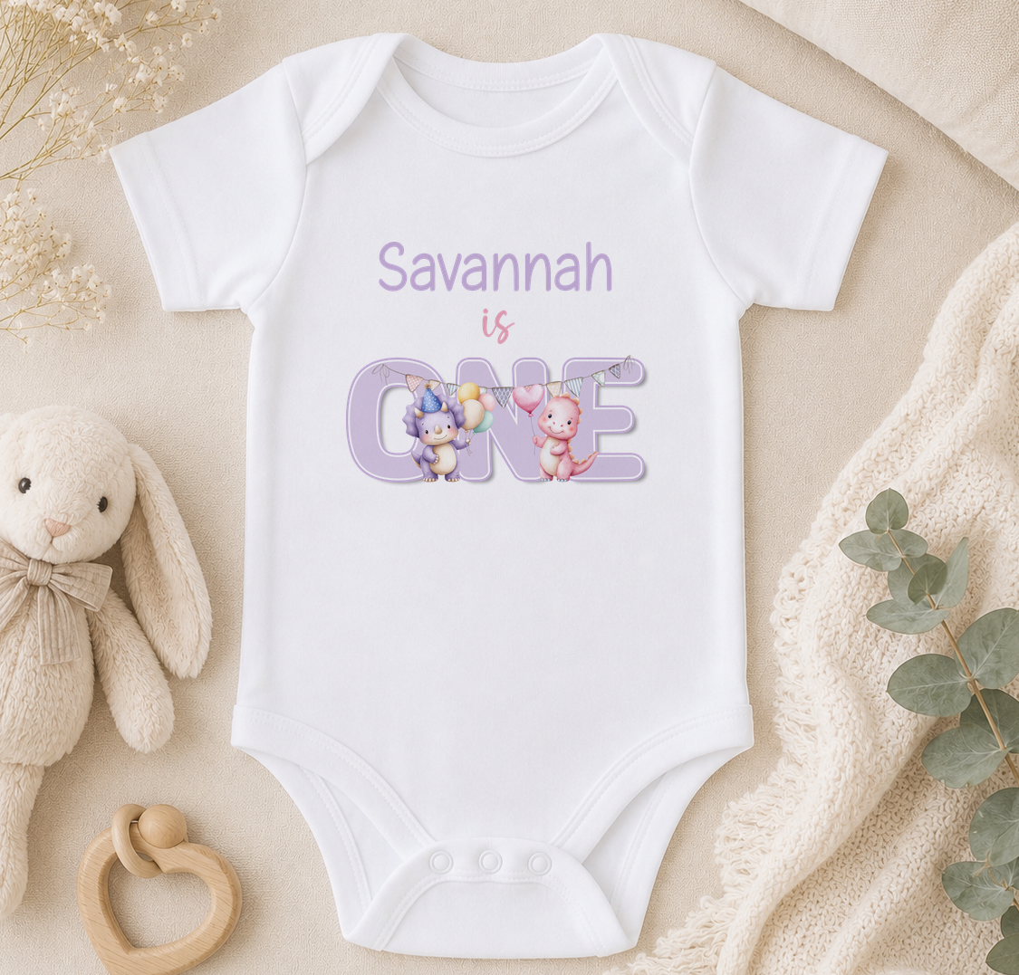A white onesie with the text "Savannah is 1" printed on it, along with illustrations of two cartoon animals. The onesie is placed on a beige surface, surrounded by a stuffed animal, a wooden toy, and some greenery.