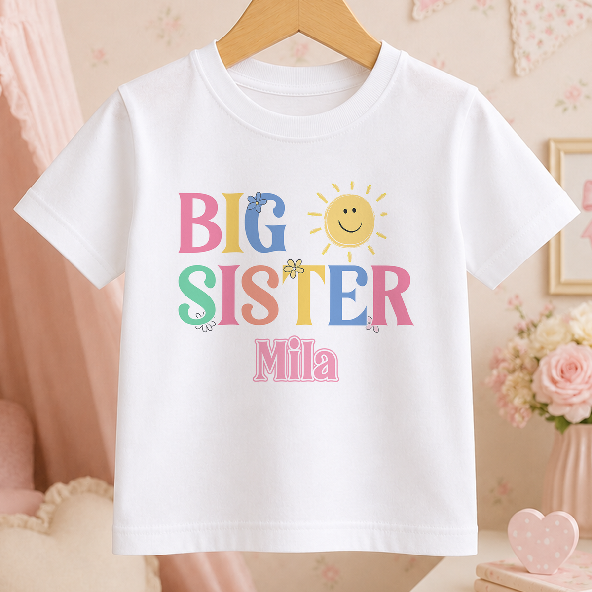 A white t-shirt with the text "BIG SISTER Mila" printed on it, along with a sun and flower design.