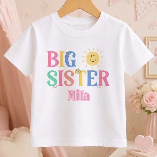 A white t-shirt with the text "BIG SISTER Mila" printed on it, along with a sun and flower design.