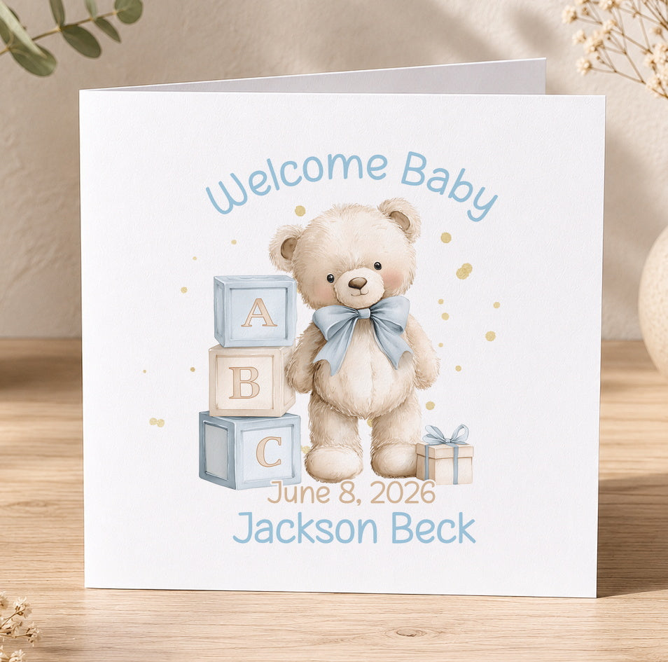 Baby congratulations card featuring a teddy bear and alphabet blocks, printed with 'Welcome Baby' and personalised name and date text.
