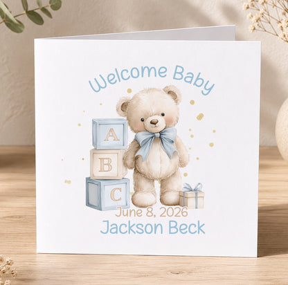 Baby congratulations card featuring a teddy bear and alphabet blocks, printed with 'Welcome Baby' and personalised name and date text.