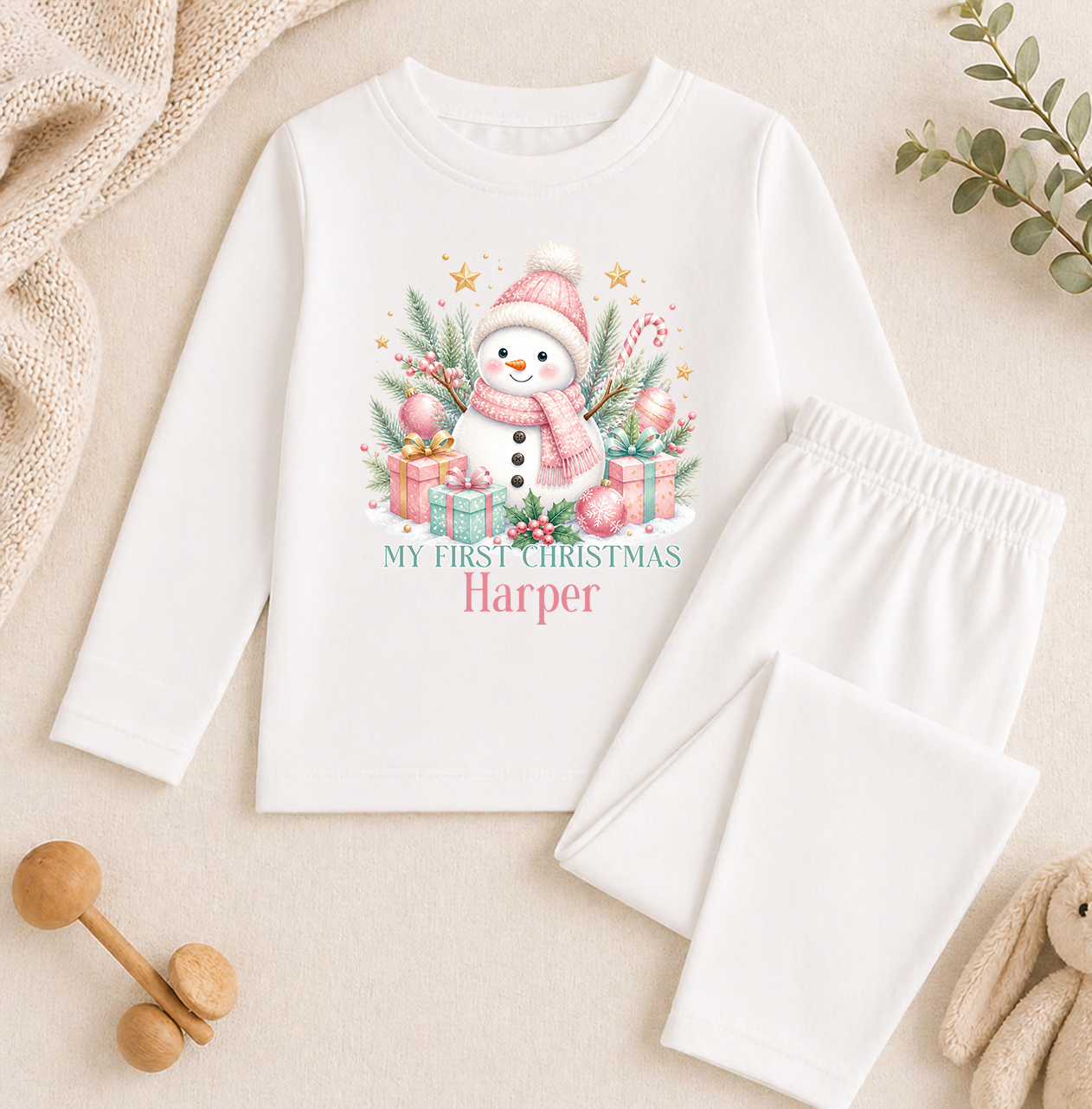 A white long-sleeved shirt with a snowman graphic and the text "My first christmas Harper" printed on it, along with a pair of white pajama pants. The shirt and pants are placed on a beige surface, and there are some toys and greenery in the background.