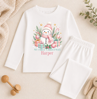 A white long-sleeved shirt with a snowman graphic and the text "My first christmas Harper" printed on it, along with a pair of white pajama pants. The shirt and pants are placed on a beige surface, and there are some toys and greenery in the background.