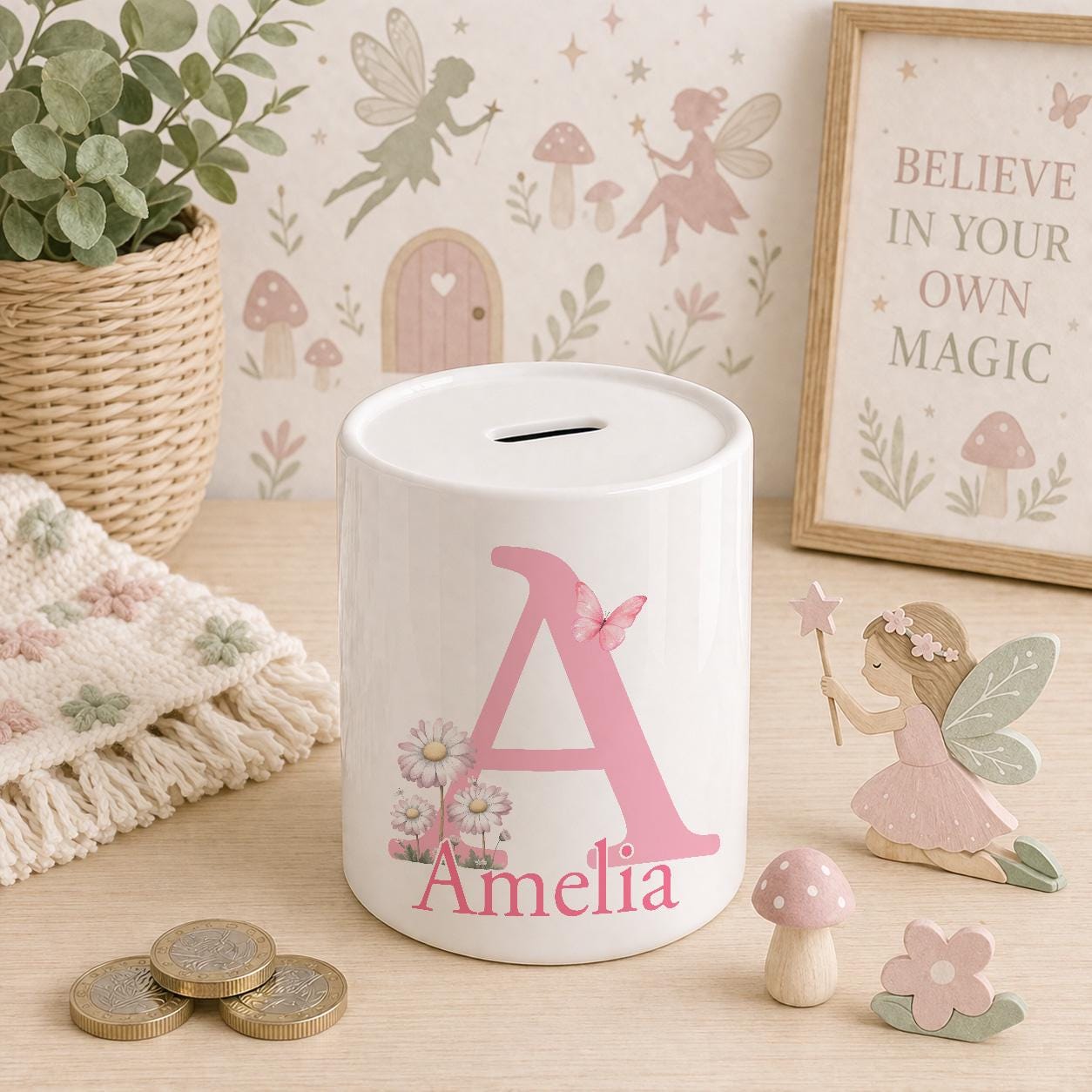 White ceramic money box personalised with the name Amelia, printed with a large pink initial letter A, a butterfly and pink daisies, displayed against a pink fairy garden background