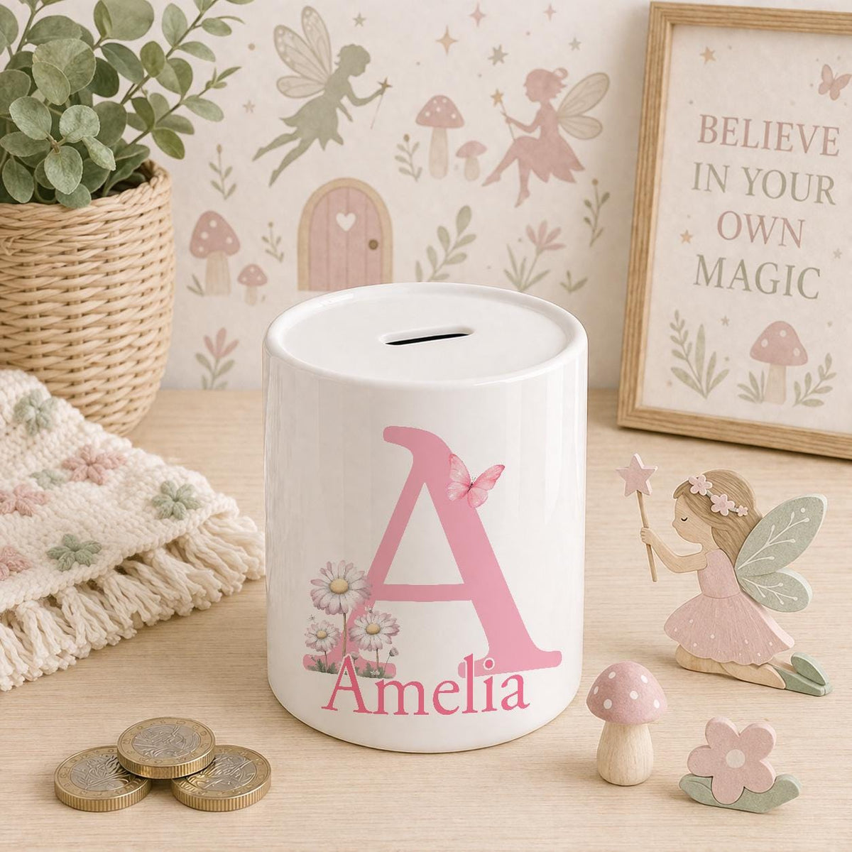 White ceramic money box personalised with the name Amelia, printed with a large pink initial letter A, a butterfly and pink daisies, displayed against a pink fairy garden background