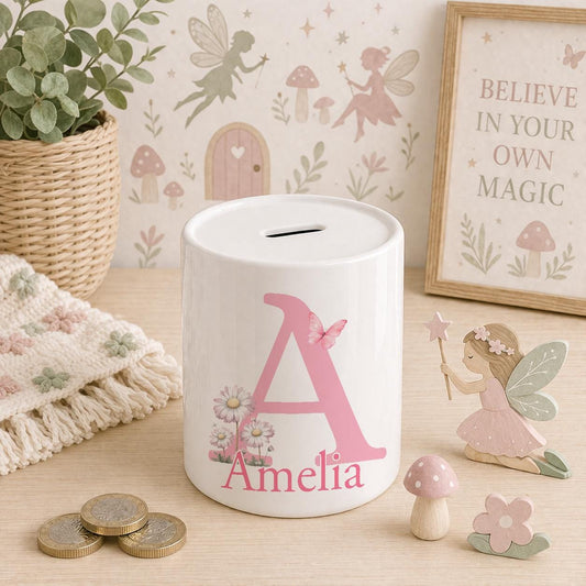 White ceramic money box personalised with the name Amelia, printed with a large pink initial letter A, a butterfly and pink daisies, displayed against a pink fairy garden background