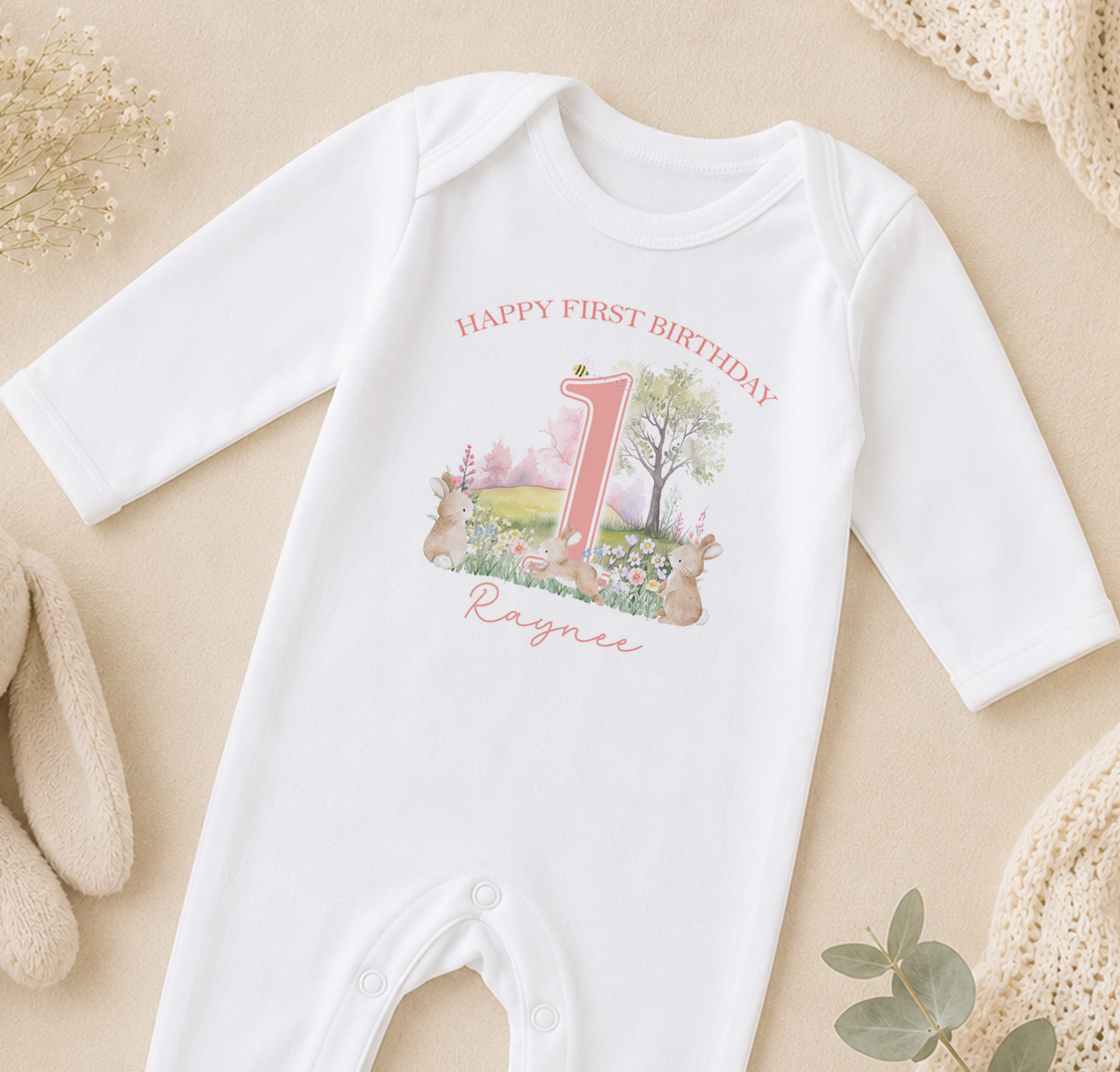 A white onesie with a pink number one and a garden scene printed on it, along with the text "Happy first birthday" and the name "Raznie".