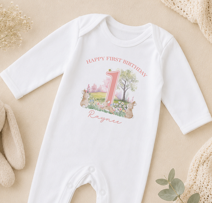 A white onesie with a pink number one and a garden scene printed on it, along with the text "Happy first birthday" and the name "Raznie".