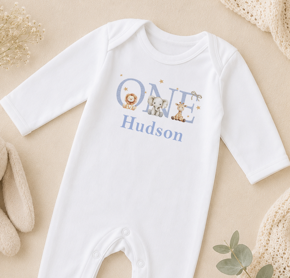 A white onesie with the name "Hudson" printed on it, along with illustrations of a lion, elephant, and giraffe.