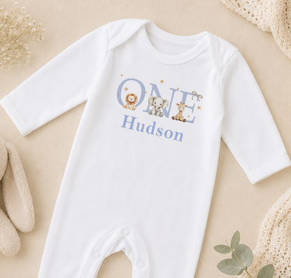 A white onesie with the name "Hudson" printed on it, along with illustrations of a lion, elephant, and giraffe.