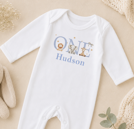A white onesie with the name "Hudson" printed on it, along with illustrations of a lion, elephant, and giraffe.