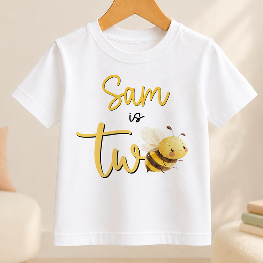 A white t-shirt with a yellow bee graphic and the text "Sam is two" printed on it.