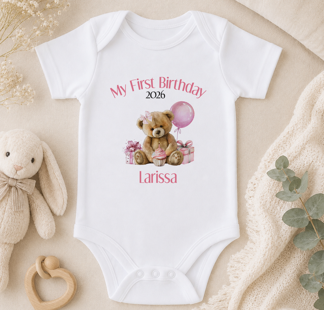 A white onesie with a teddy bear and a balloon, along with the text "My First Birthday 2026 Larissa" printed on it, is displayed on a bed with a stuffed animal and a blanket.