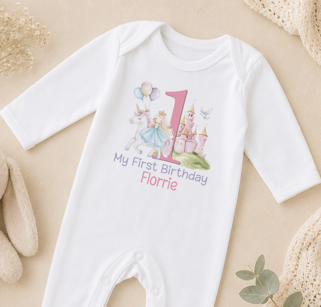 A white onesie with a personalized birthday message and a unicorn graphic, along with a castle and the number 1, is displayed on a beige surface.