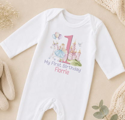 A white onesie with a personalized birthday message and a unicorn graphic, along with a castle and the number 1, is displayed on a beige surface.