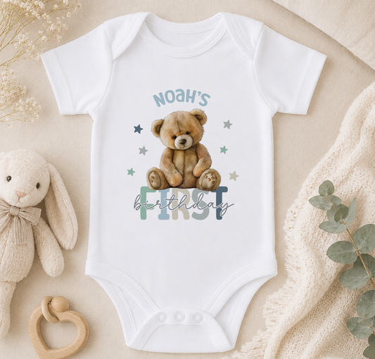A white baby onesie with a teddy bear and the text "Noah's first birthday" printed on it, along with a teddy bear and other baby items.