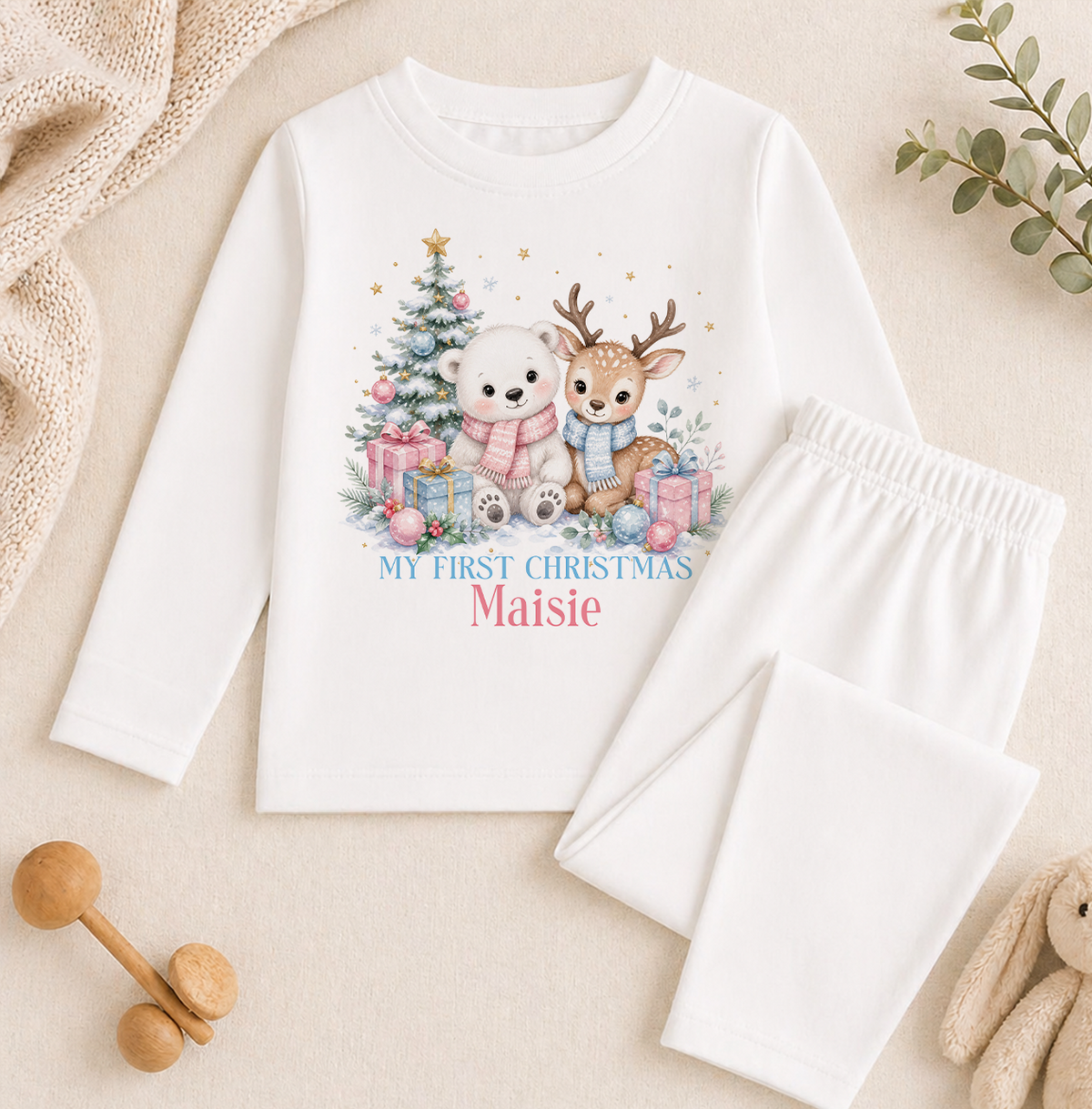 A white long-sleeved shirt with a Christmas-themed graphic and text, along with a pair of white pajama pants, and some stuffed animals, all laid out on a beige background.