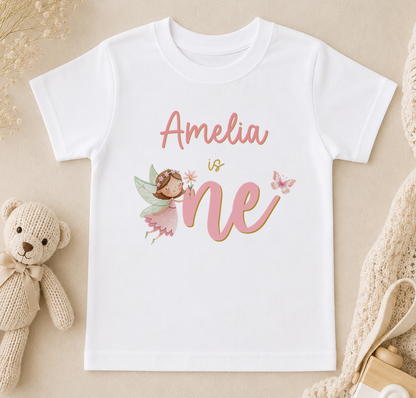 A white t-shirt with a personalized text and a fairy graphic, along with a teddy bear and other baby items, arranged on a beige surface.