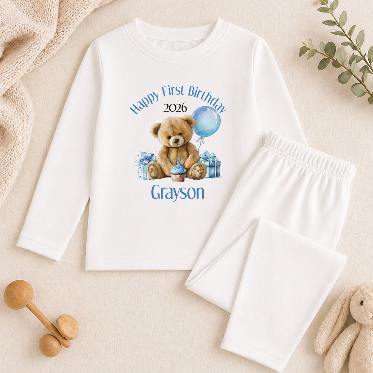 A white long-sleeved shirt with a teddy bear graphic and text, along with a pair of white pants, is displayed on a beige surface. There are also some stuffed animals and a plant in the background.