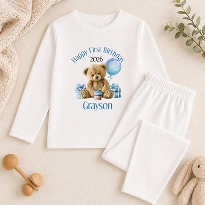 A white long-sleeved shirt with a teddy bear graphic and text, along with a pair of white pants, is displayed on a beige surface. There are also some stuffed animals and a plant in the background.