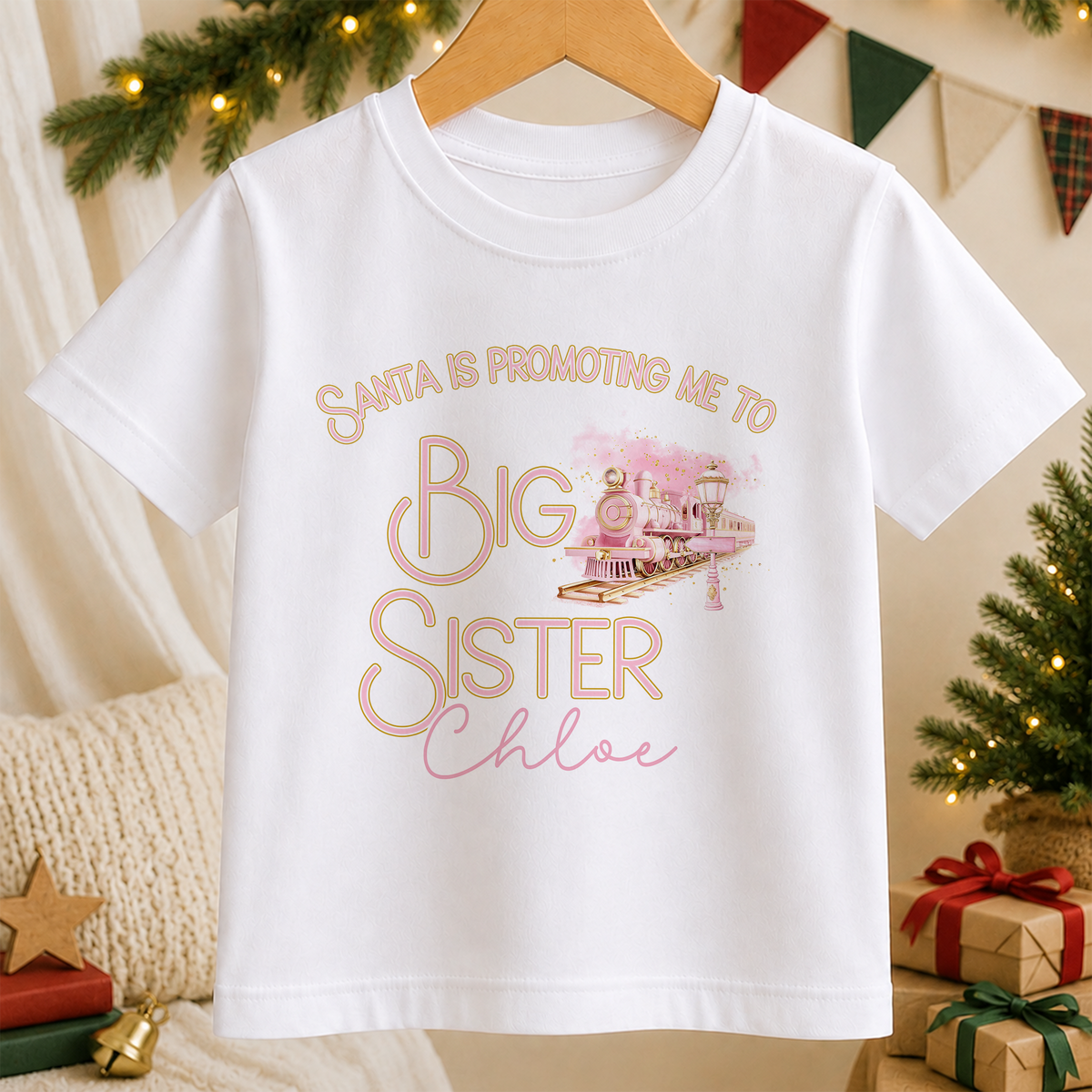 A white children's t-shirt printed with a pink express train design and the words 'Promoted to Big Sister', personalised with a child's name, hanging on a wooden hanger.