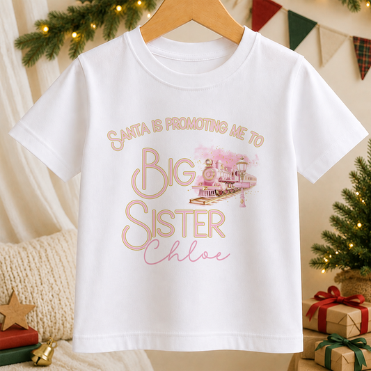 A white children's t-shirt printed with a pink express train design and the words 'Promoted to Big Sister', personalised with a child's name, hanging on a wooden hanger.