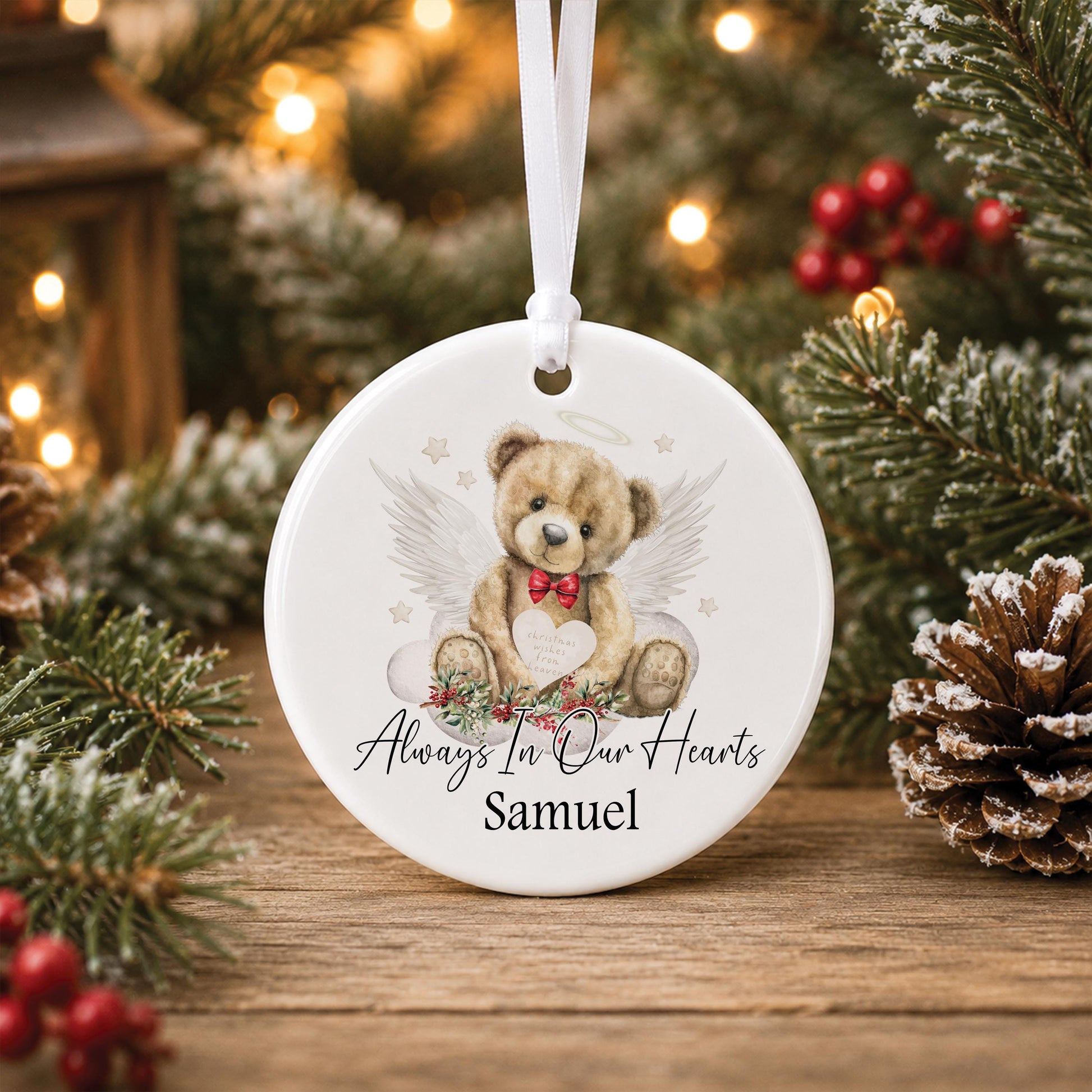 Personalised round ceramic memorial Christmas bauble featuring a watercolour angel teddy bear with white wings, red bow tie, festive foliage and Always In Our Hearts wording.