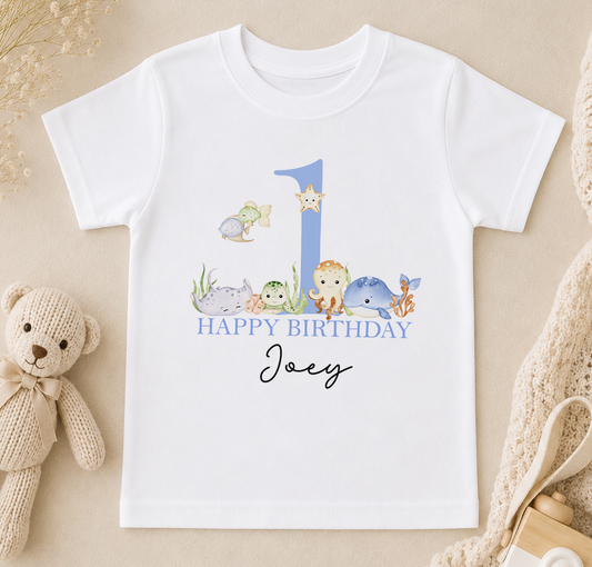 A white t-shirt with a graphic design featuring a blue number 1, surrounded by various sea animals, and the text "Happy Birthday Joey" printed on it.