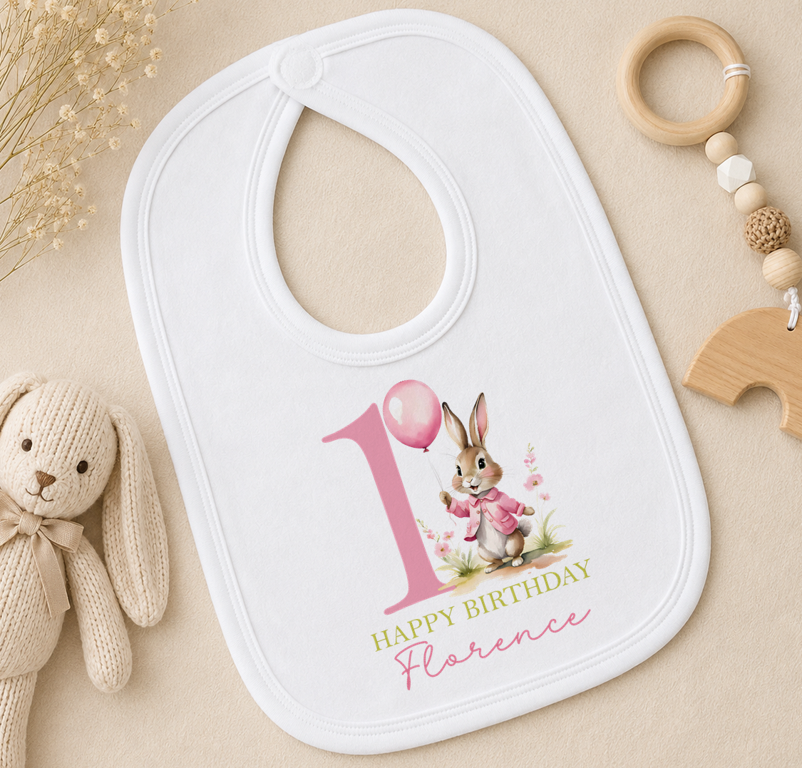 A white bib with a pink bunny and the number 1, along with the text "Happy Birthday Florence" printed on it, is placed on a surface next to a stuffed rabbit and a wooden bead ringer.