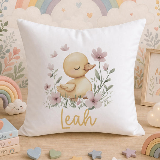 A white cushion printed with a watercolour duckling and blush-pink flowers, personalised with a child's name, photographed on a wooden surface with a colourful rainbow-themed background.
