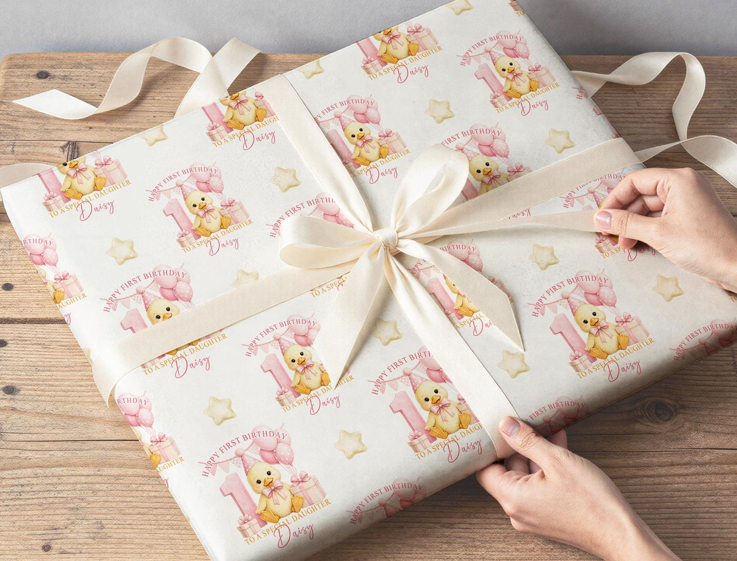 Personalised first birthday wrapping paper wrapped around a gift with cream ribbon, featuring a cute duckling, pink number one, balloons and presents.