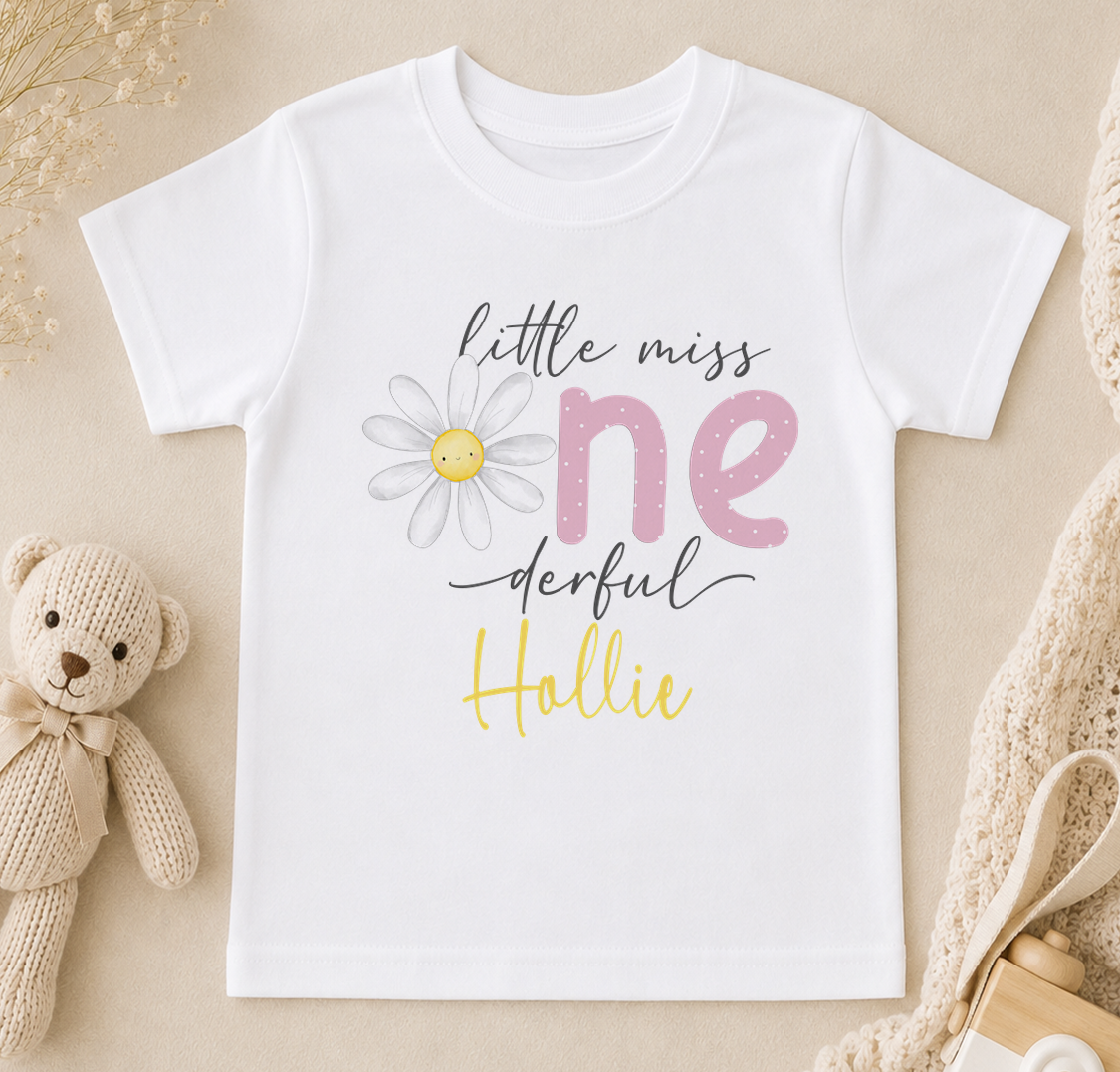 A white t-shirt with a flower design and text printed on it, placed next to a stuffed teddy bear and some other items.