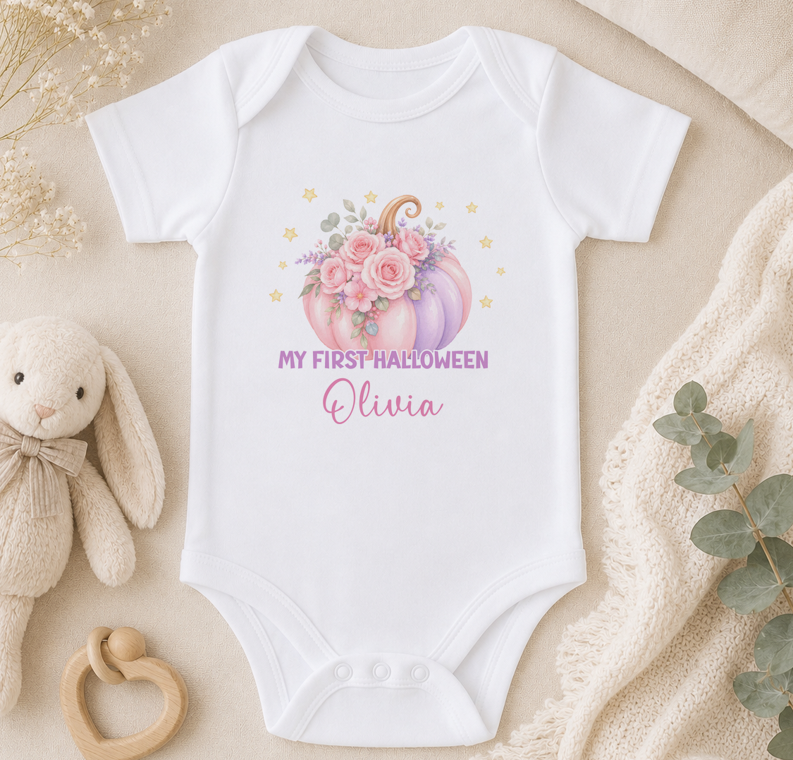 A white baby onesie with a pumpkin and flowers design, along with a stuffed bunny and a beige blanket, arranged on a surface.