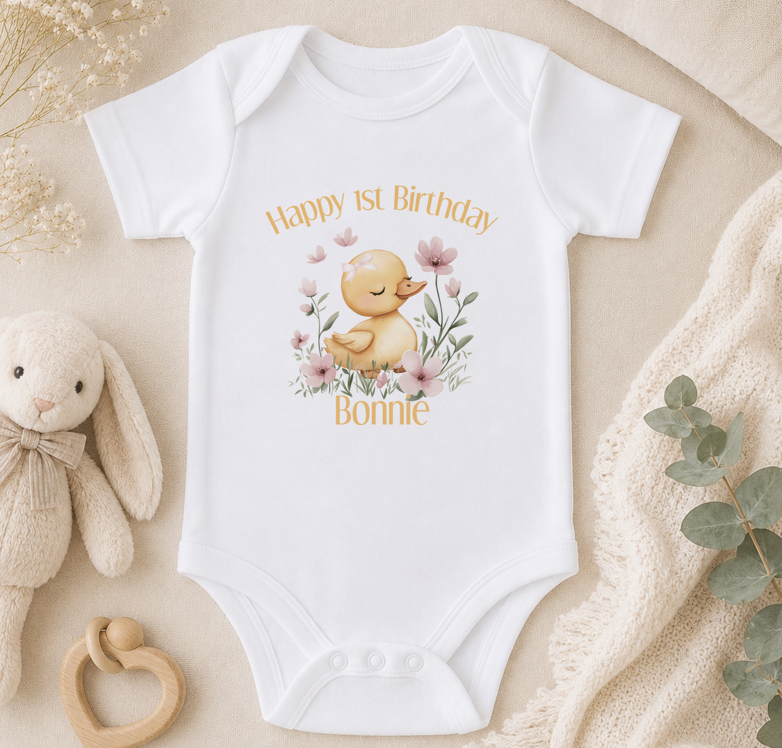 A white onesie with a cartoon duck and flowers, along with a stuffed bunny and a beige blanket, arranged on a surface.