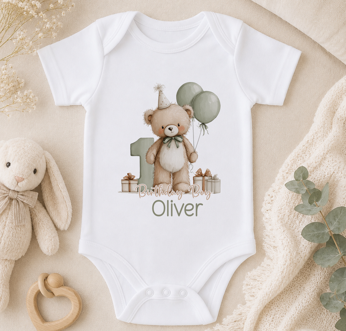 A white onesie with a teddy bear and balloons, along with the text "Birthday Boy Oliver" printed on it, is displayed on a bed with a stuffed bunny and some greenery.