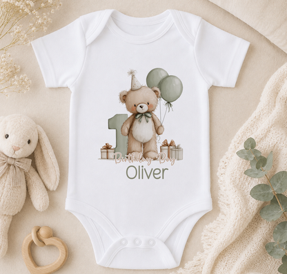 A white onesie with a teddy bear and balloons, along with the text "Birthday Boy Oliver" printed on it, is displayed on a bed with a stuffed bunny and some greenery.