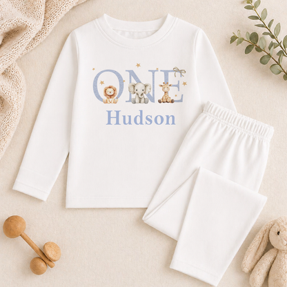 A white onesie with the name "Hudson" printed on it, along with a pair of white pajama pants, and some stuffed animals.