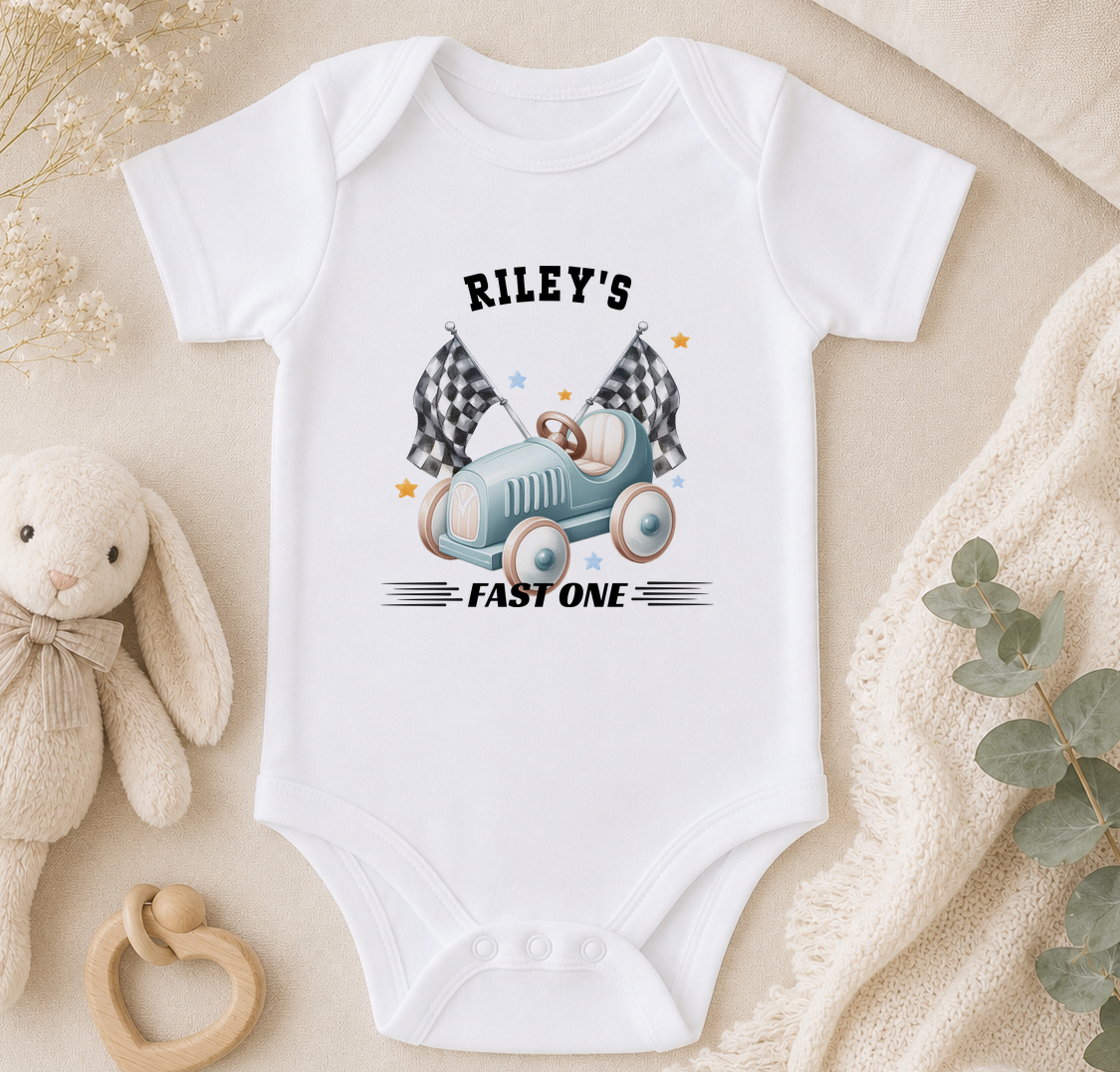 White baby onesie printed with a car and chequered flags design and the text 'Riley's FAST ONE', styled with a soft toy and rattle.