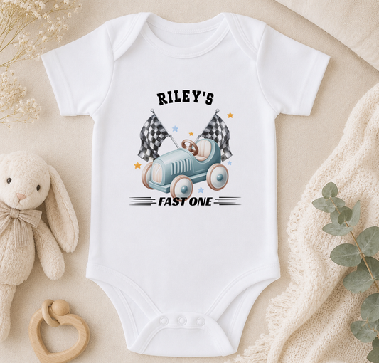 A white baby onesie with a graphic of a car and checkered flags, along with the text "Riley's FAST ONE". The onesie is placed on a beige surface, surrounded by a stuffed animal, a baby rattle, and some greenery.