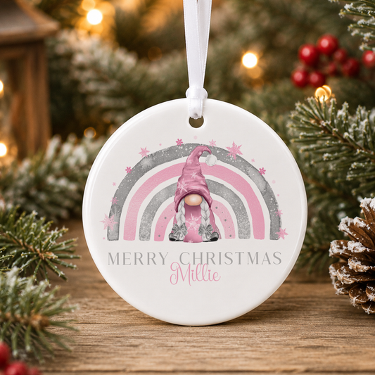 A white ornament with a pink gnome and a rainbow graphic, surrounded by snow and pine branches, with the text "MERRY CHRISTMAS MILLIE" displayed on it.