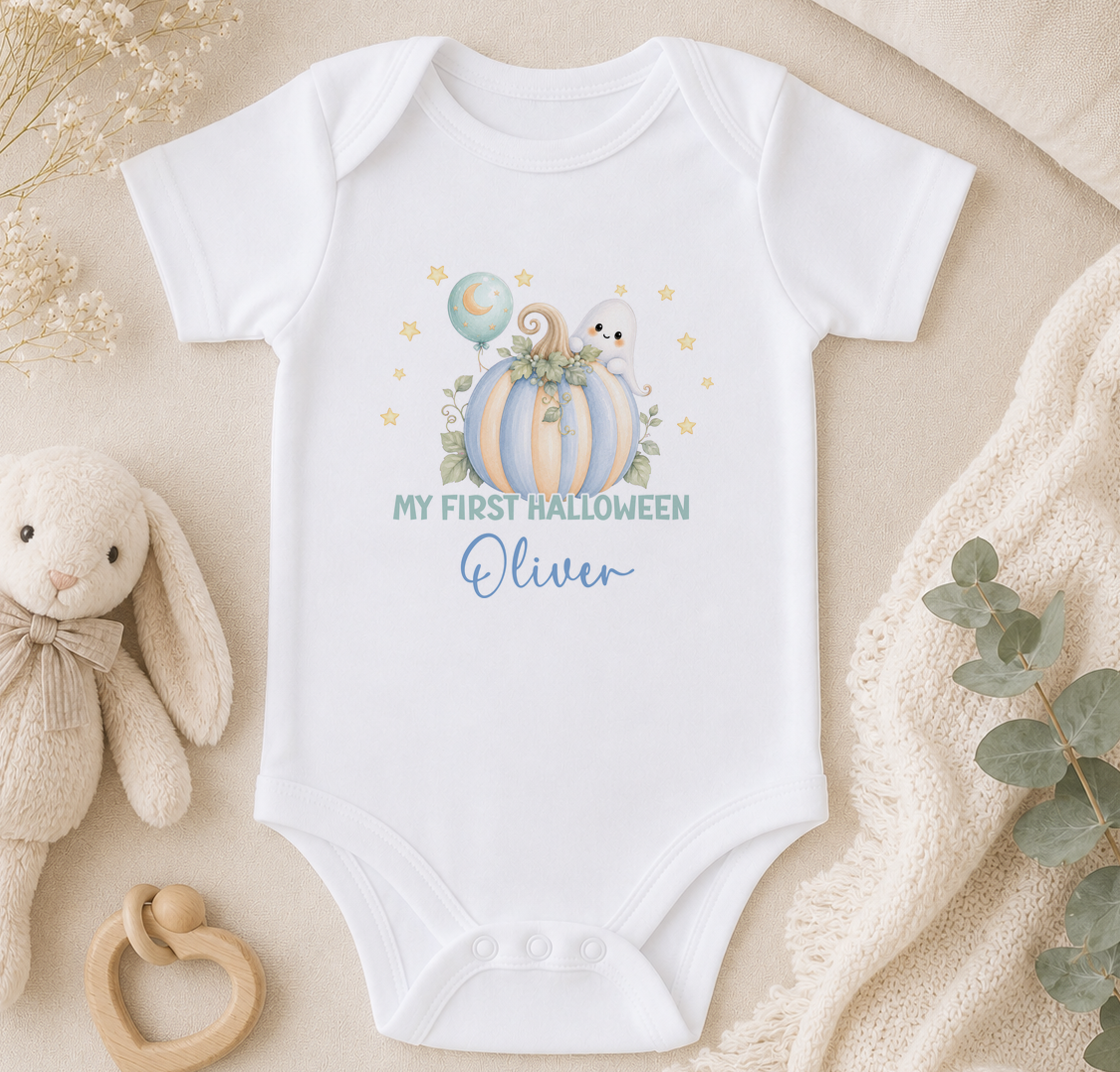 A white baby onesie with a Halloween-themed design, including a pumpkin, ghost, and stars, along with the text "My first Halloween Oliver". The onesie is placed on a beige surface, surrounded by a stuffed bunny, a wooden bead rattle, and some greenery.