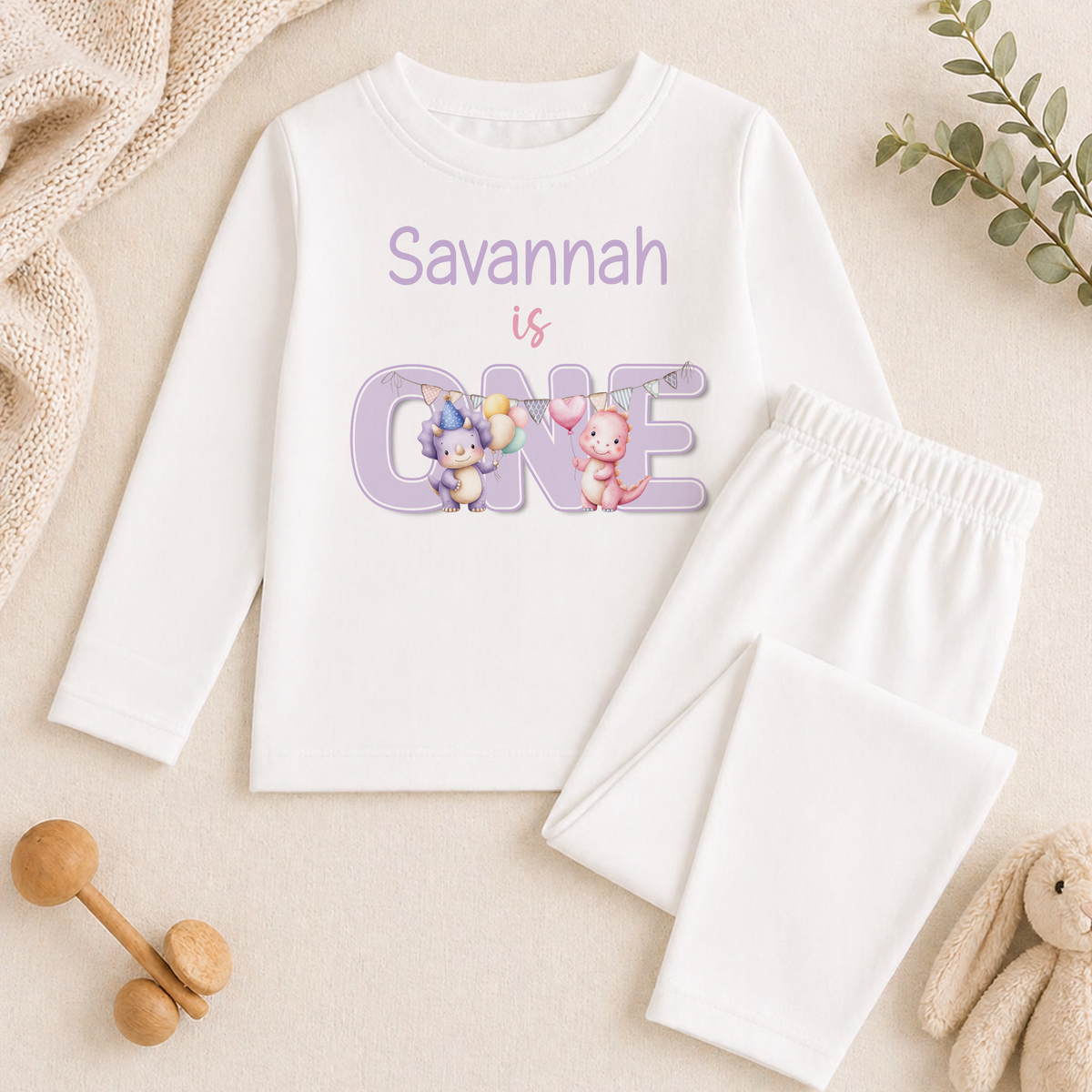 A white long-sleeved shirt with the text "Savannah is ONE" printed on it, along with illustrations of two cartoon animals, is displayed on a beige surface. Next to the shirt are a pair of white pants and some stuffed animals, creating a casual, child-friendly setting.