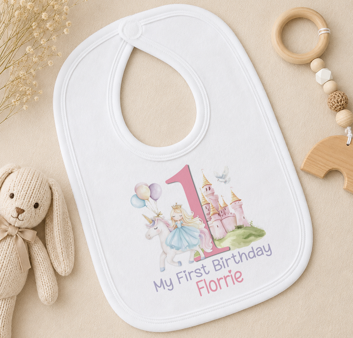 A white bib with a princess and castle design, along with the text "My First Birthday Florrie", is placed on a surface alongside a stuffed rabbit and a wooden bead ring.