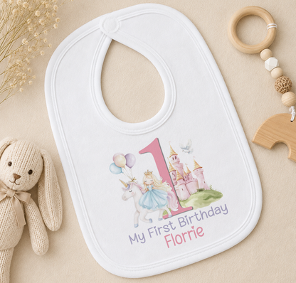 A white bib with a princess and castle design, along with the text "My First Birthday Florrie", is placed on a surface alongside a stuffed rabbit and a wooden bead ring.