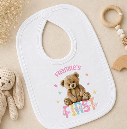 A white baby bib with a teddy bear and the text "Frankie's First Birthday" printed on it, along with a teddy bear and other baby items.
