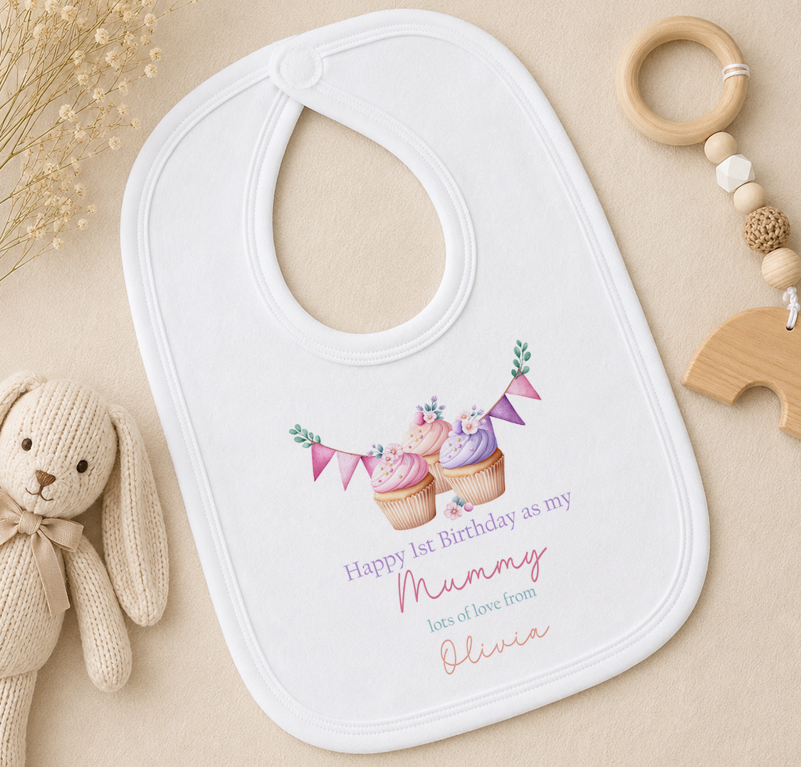 A white bib with a personalized message, a wooden toy, and some dried flowers on a beige surface.