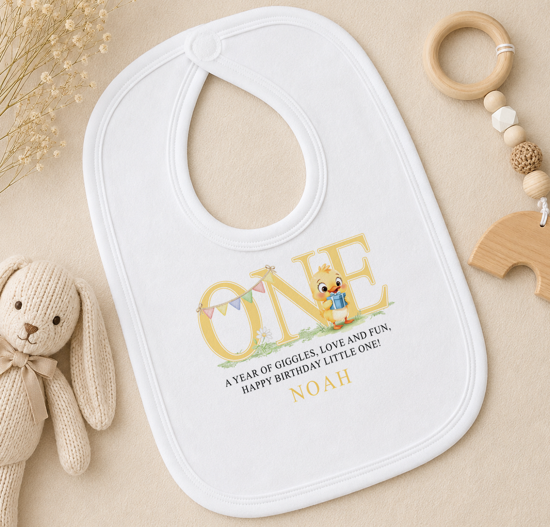 A white baby bib with a personalized message, a wooden toy, and some dried flowers on a beige surface.