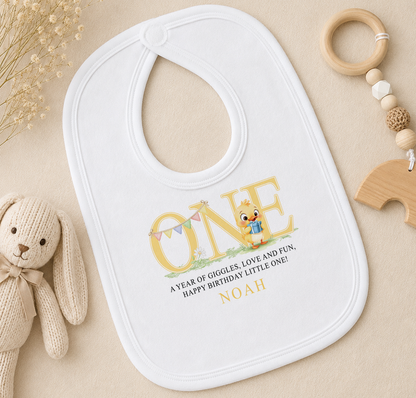 A white baby bib with a personalized message, a wooden toy, and some dried flowers on a beige surface.