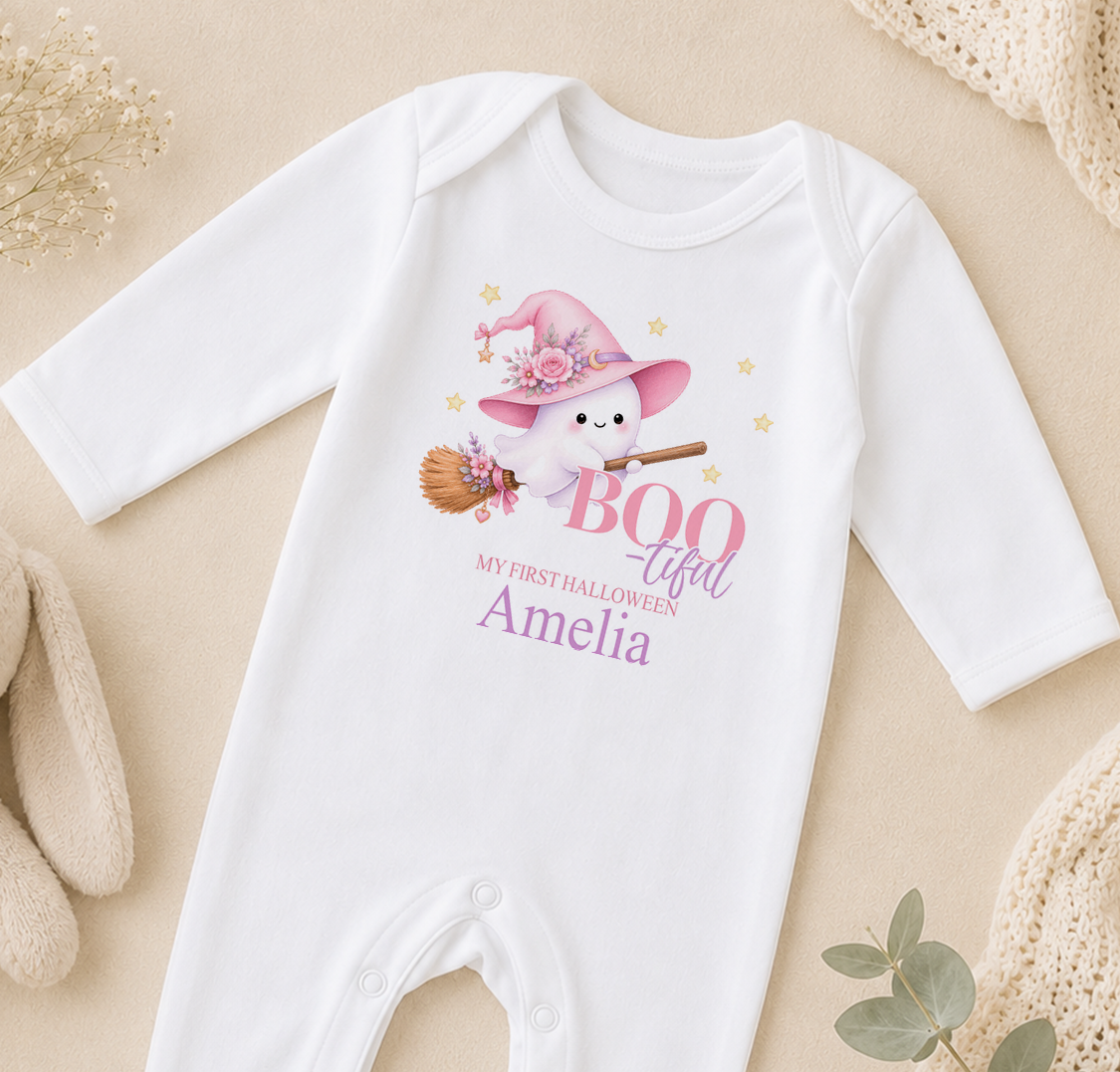 A white onesie with a cartoon ghost wearing a witch's hat and holding a broom, along with the text "My first Halloween Amelia" printed on it.