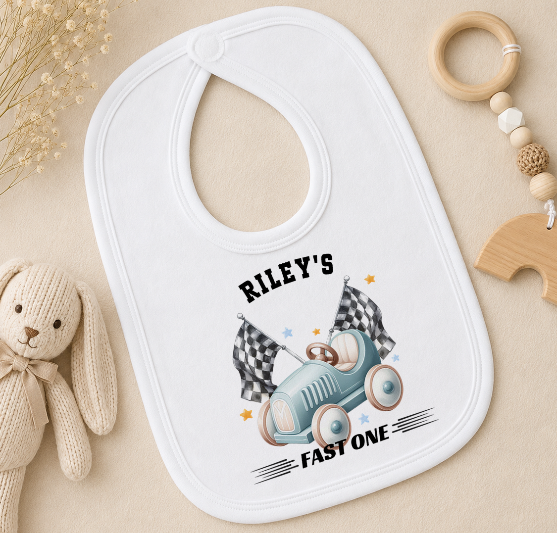 White baby bib printed with a race car and chequered flags design, styled with a soft toy and wooden ring.